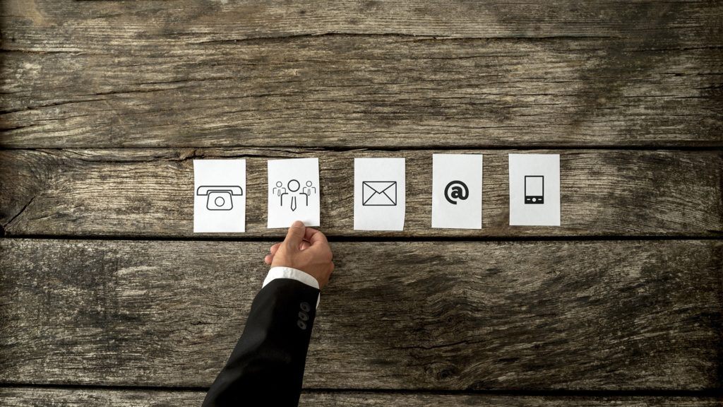 Hand selecting modern communication icons on rustic wooden table: camera, silhouettes, envelope, @ symbol, smartphone. Managed Services.