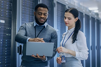 two people standing in front of servers looking at a laptop