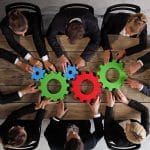 Collaborative professionals brainstorming synergistic ideas around colorful gears in teamwork meeting.