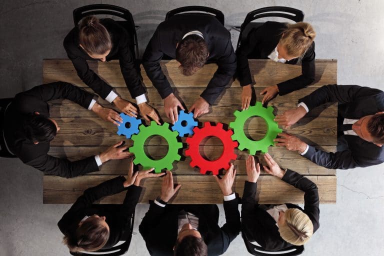 Collaborative professionals brainstorming synergistic ideas around colorful gears in teamwork meeting.