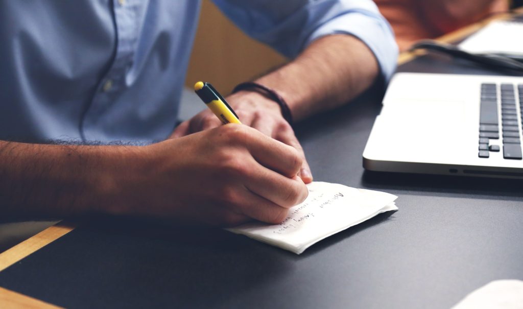 person writing on notepad with yellow pen in modern workspace, blending traditional and digital tools.