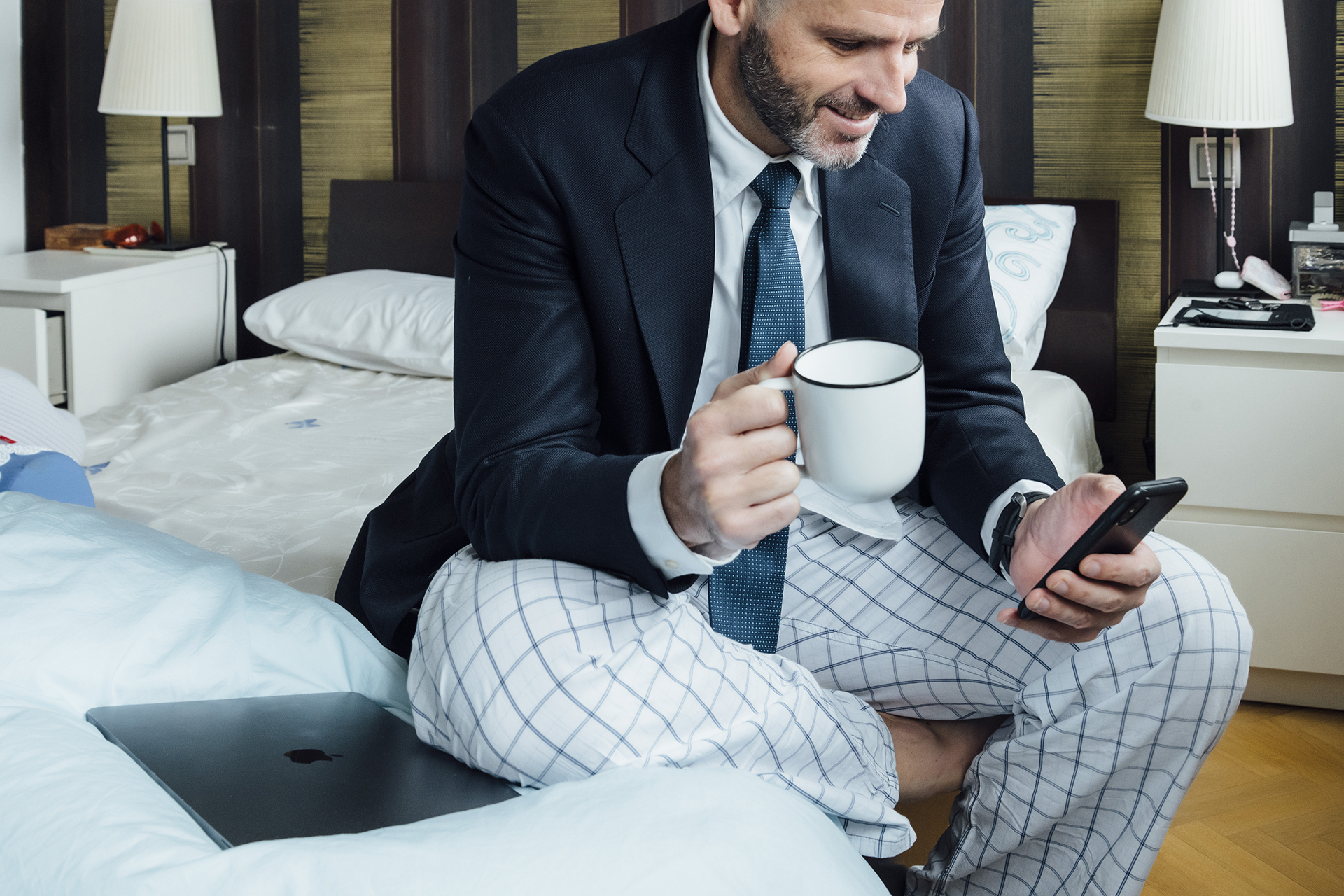 a man sitting on a bed holding a cup and cell phone