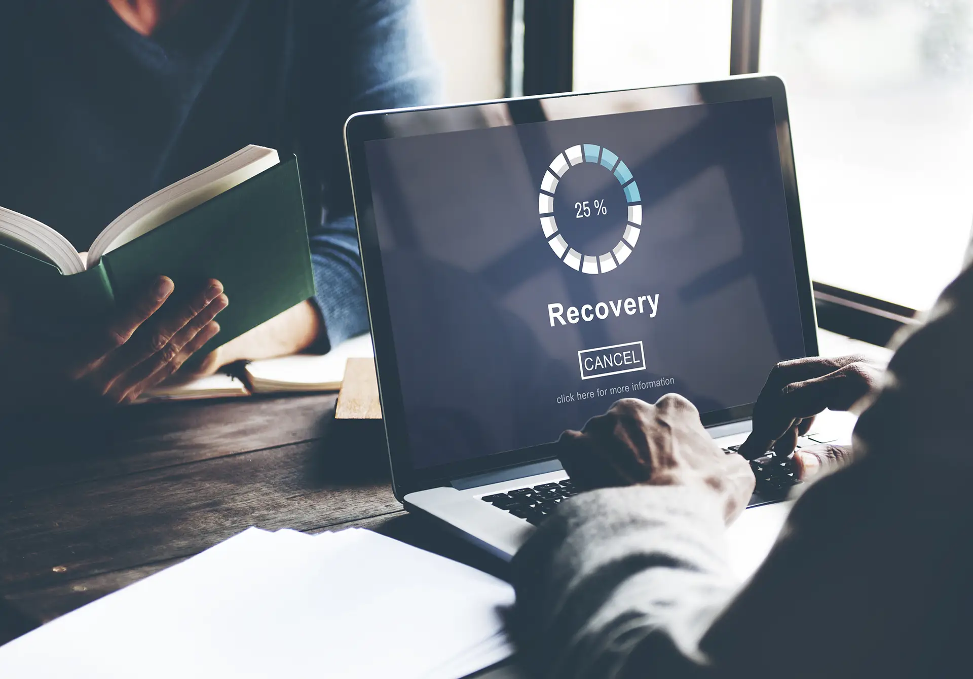 Tech recovery process on laptop screen while person reads book in cozy workspace.