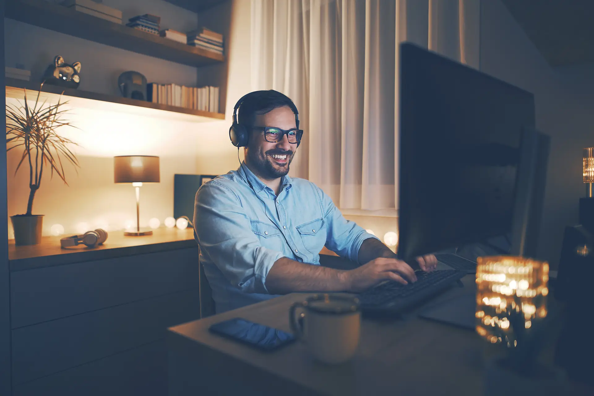 Cozy home office setup with man smiling at laptop in softly lit environment.
