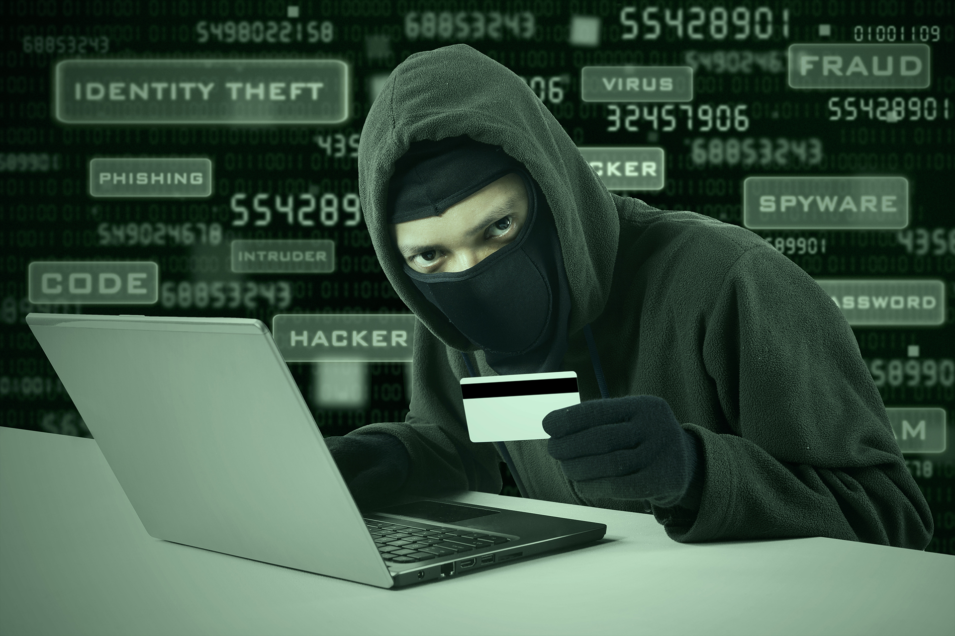 a person wearing a mask and holding a credit card in front of a laptop