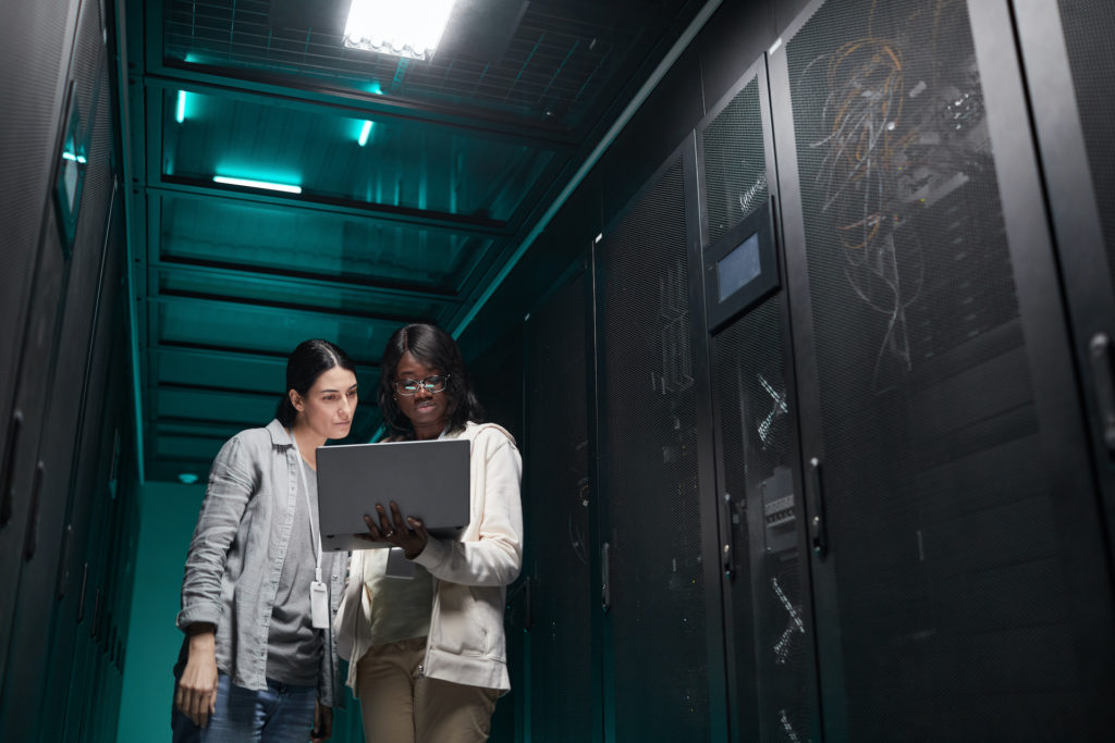 two women standing in a server room looking at a laptop