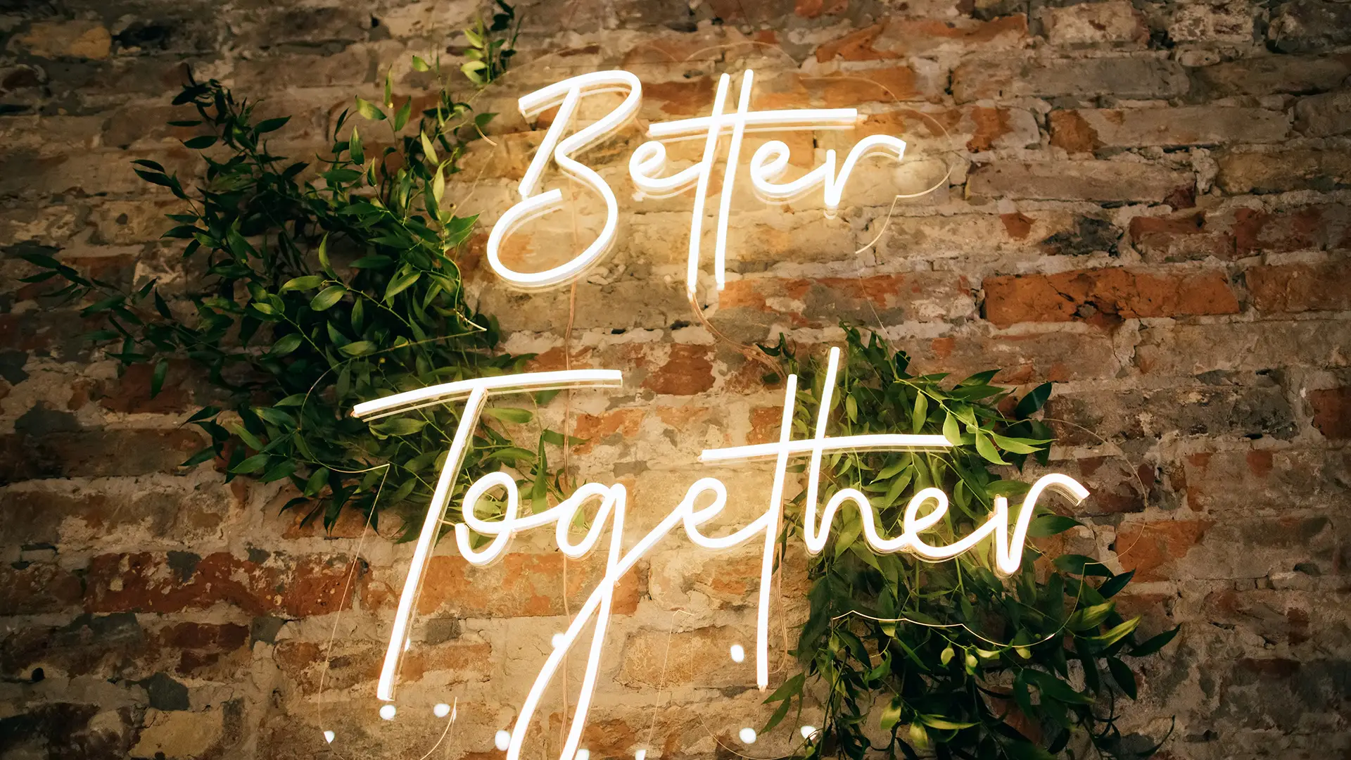 Glowing neon sign Better Together on rustic brick wall with green foliage.
