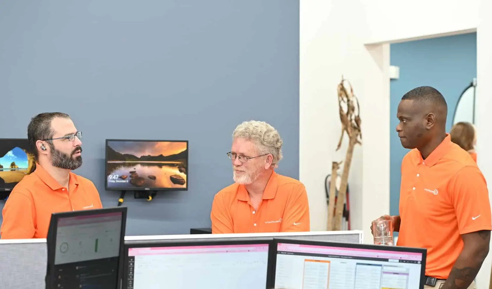 three men in orange polo shirts having a friendly conversation in a modern workplace.