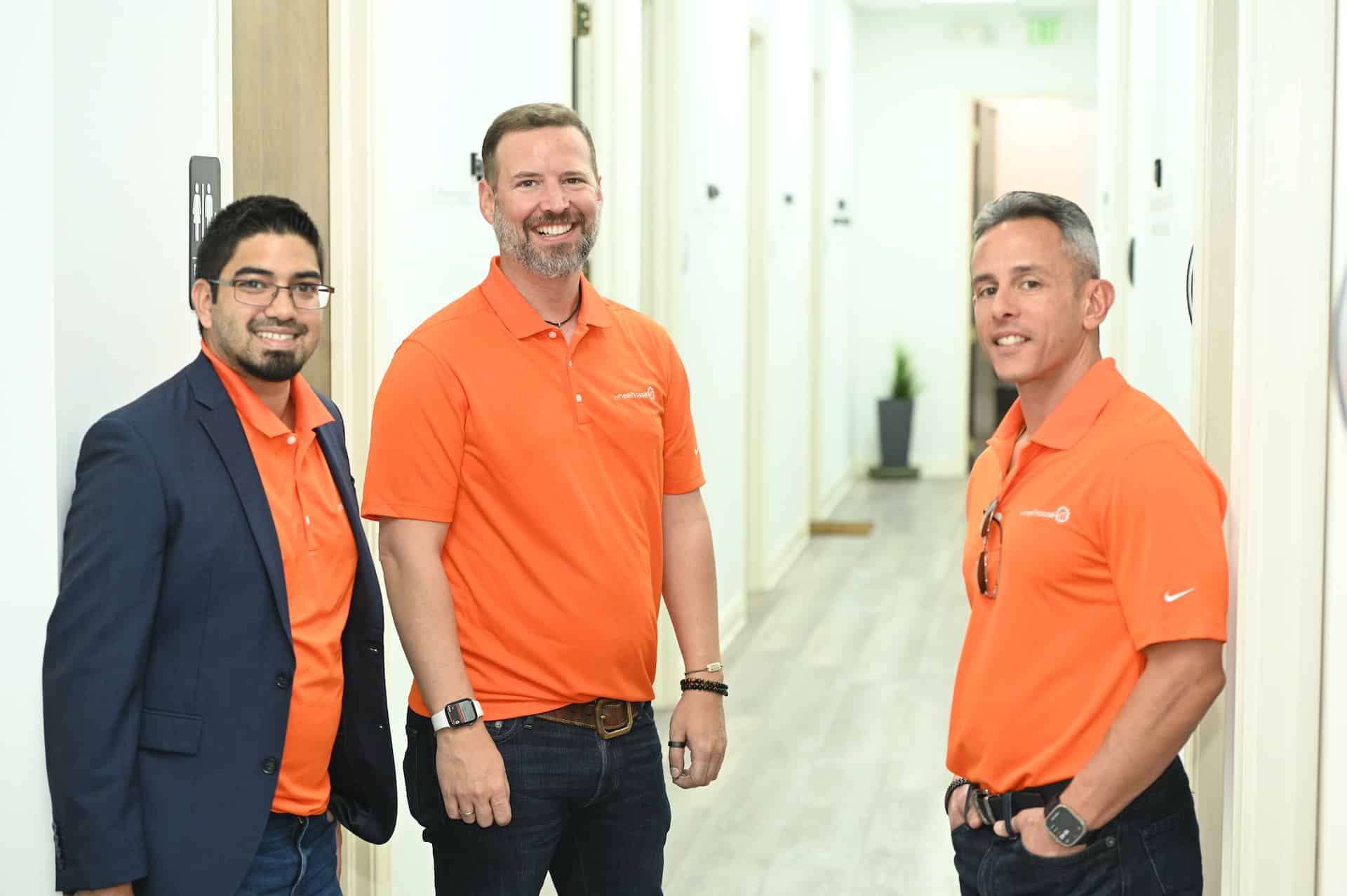 three professionals in orange polo shirts standing in a well-lit hallway.