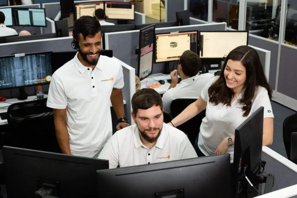 three office coworkers collaborate at a computer workstation in a modern cubicle workspace.