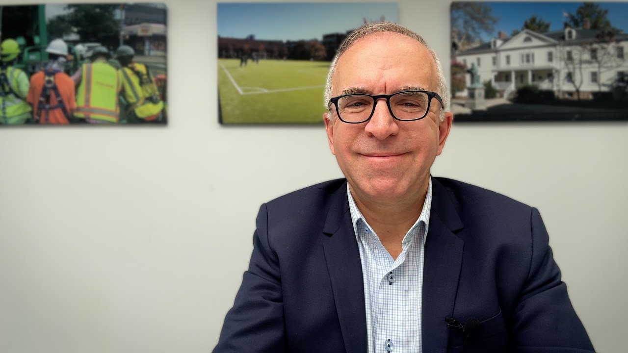 middle-aged man in professional attire smiling at desk with community themed background photos.