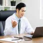 Asian male doctor conducts a telemedicine consultation by laptop in a bright medical office.