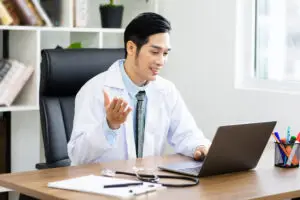 asian male doctor conducts a telemedicine consultation by laptop in a bright medical office.